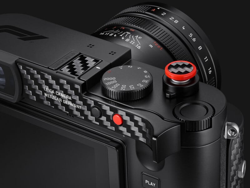 A close-up view of a black camera with carbon fiber accents, featuring a shutter speed dial, a red shutter button, and a lens with aperture markings.