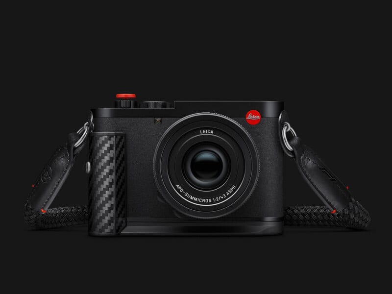 A black Leica camera with a carbon fiber grip, a red shutter button, and a black braided strap against a dark background.