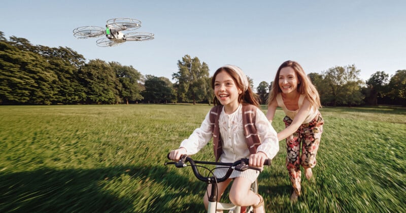A woman runs behind a young girl riding a bicycle through a grassy field while a small drone flies nearby.
