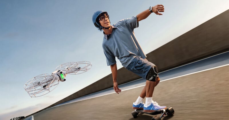 A person wearing a helmet and glasses rides an electric skateboard while controlling a small, white drone nearby.