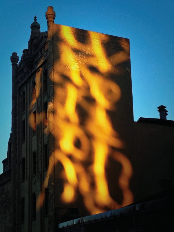 Golden sunlight reflects off a nearby surface, casting abstract, glowing patterns onto the side of a tall, historic brick building against a blue sky.