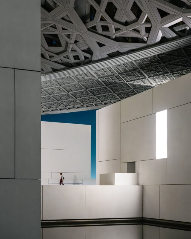 A person walks along a white walkway beneath a large, intricate geometric dome structure at the Louvre Abu Dhabi.
