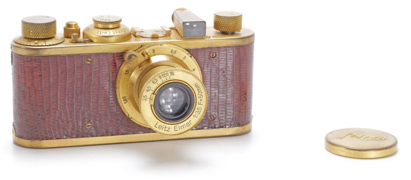 A gold-toned vintage camera with a brown textured body and a Leitz Elmar 50mm lens, accompanied by a matching lens cap on a white background.