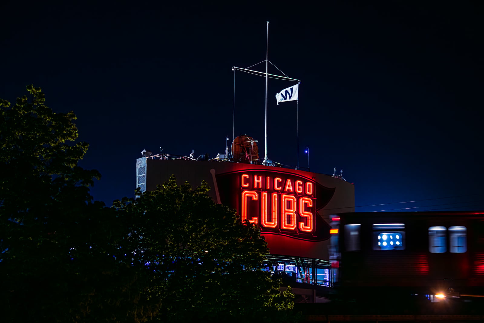 A neon "CHICAGO CUBS" sign glows at night above a stadium, with a "W" flag flying on a pole above it and a blurred train passing in the foreground.