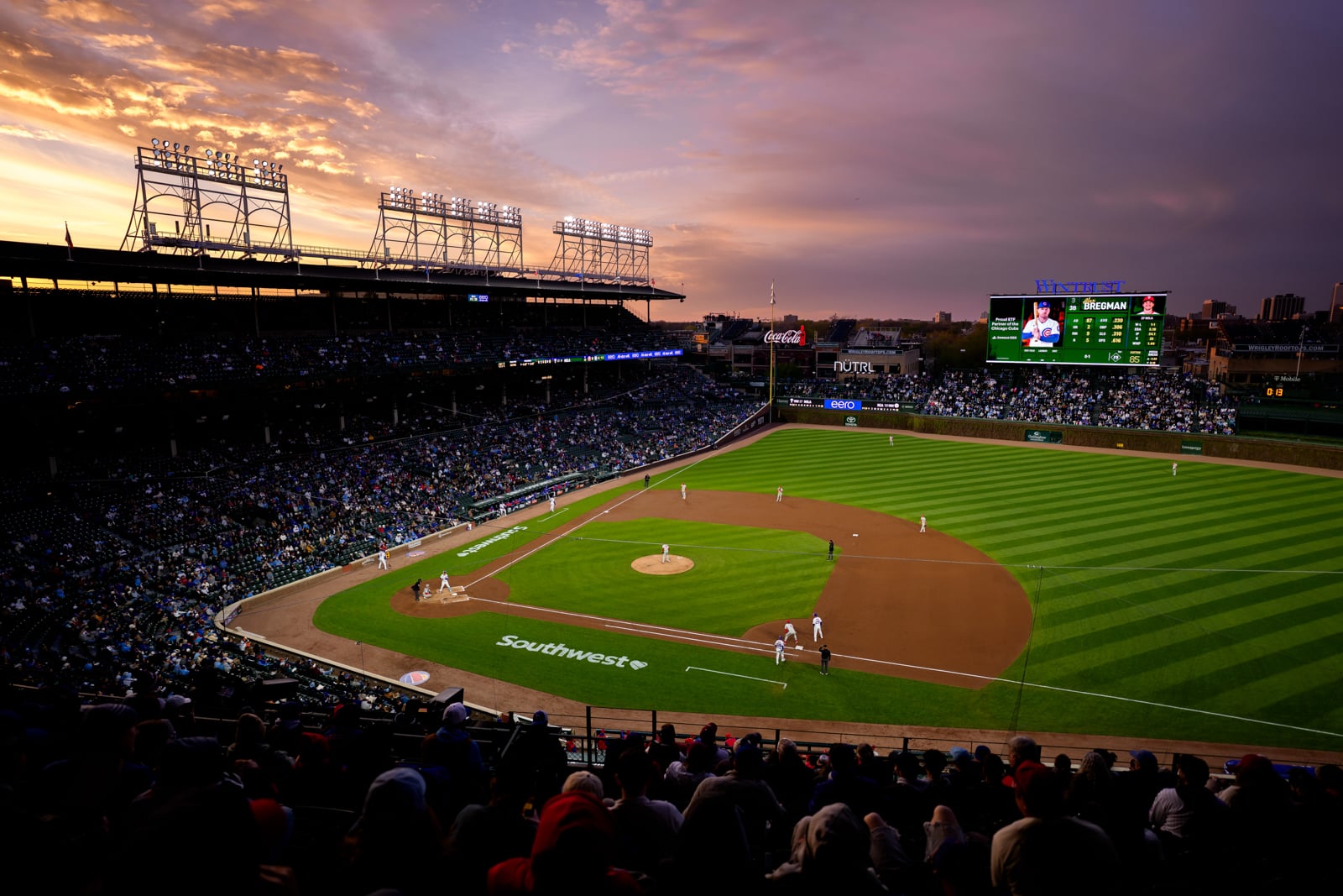A wide-angle view of a baseball stadium at sunset, with players on the field and a large digital scoreboard displaying player statistics.