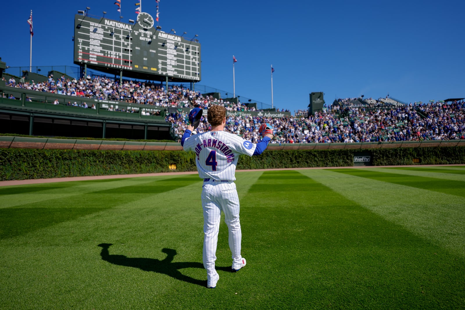 A baseball player wearing a white pinstriped jersey with the name "CROW-ARMSTRONG" and number 4 stands on the field at Wrigley Field, facing the scoreboard with his hat raised.