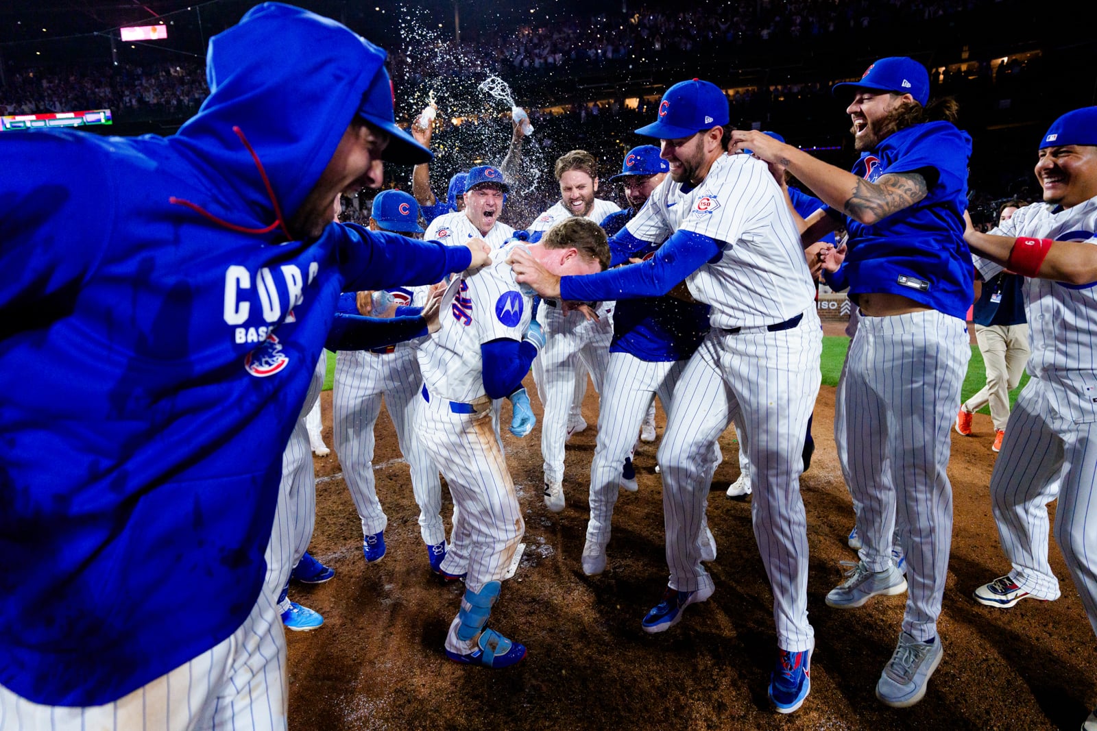 Chicago Cubs baseball players celebrate on the field, dousing a teammate with water while wearing team uniforms and hoodies.