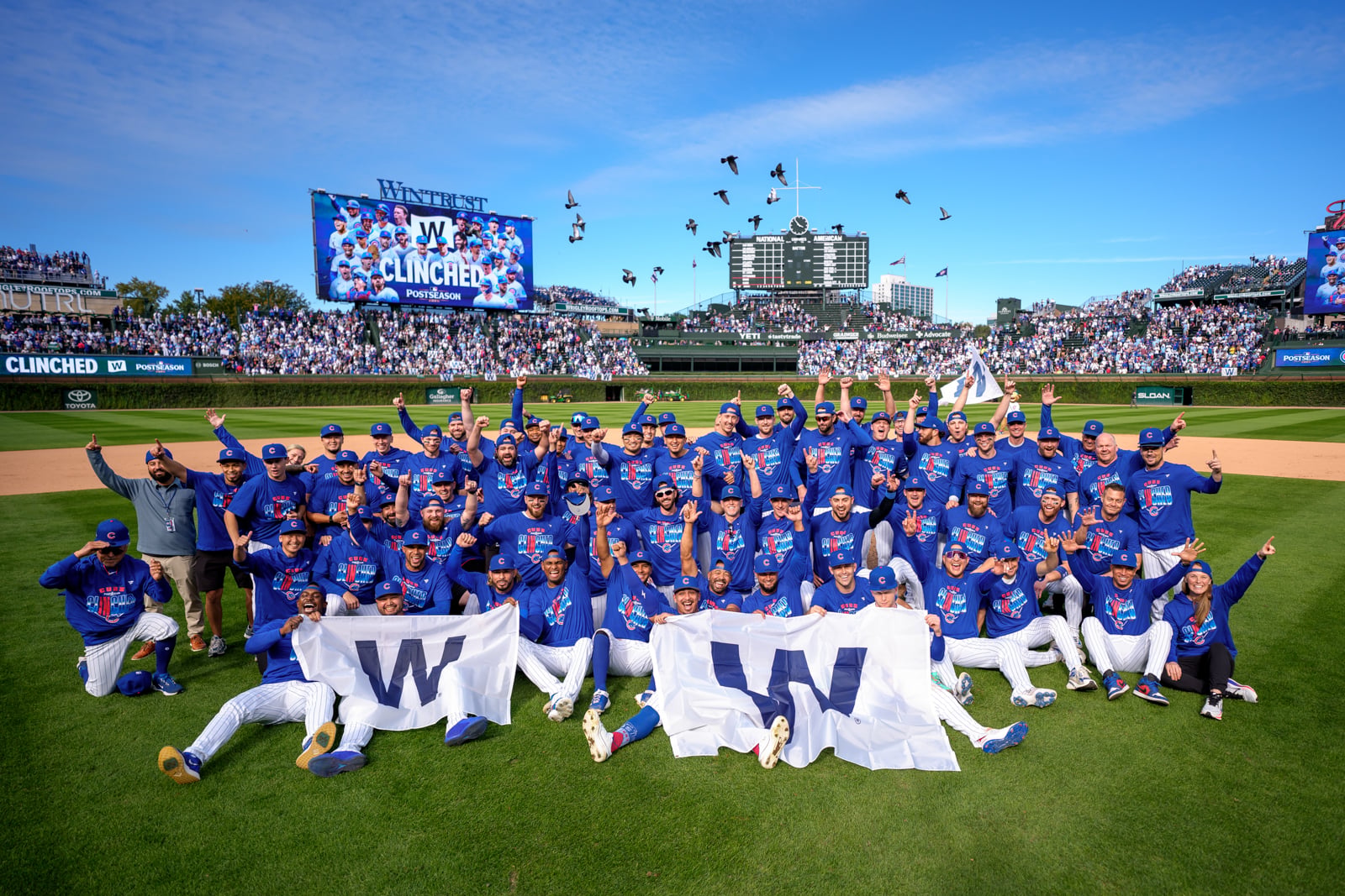 The Chicago Cubs baseball team celebrates on the field at Wrigley Field after clinching a postseason spot, holding large "W" flags.