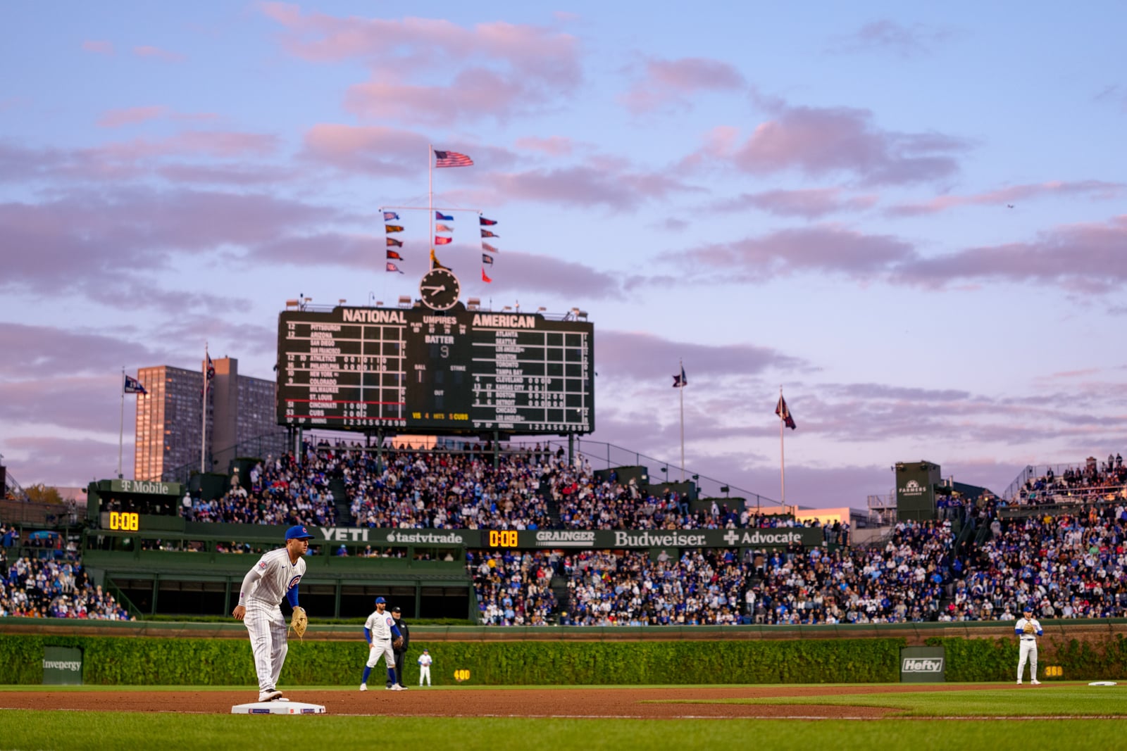 A Chicago Cubs baseball player stands on first base during a game at Wrigley Field, with the iconic manual scoreboard and a crowd of spectators in the background under a sunset sky.