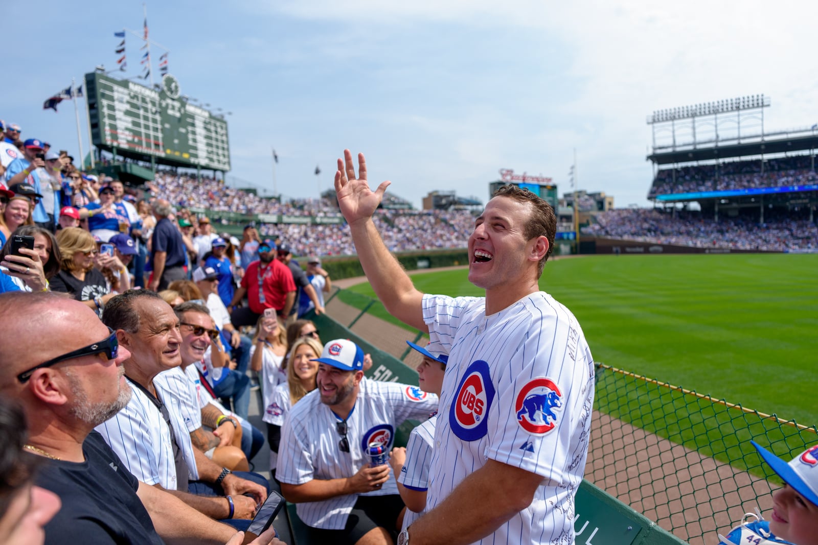 A man in a Chicago Cubs jersey waves to a crowd from behind a stadium fence while other fans watch from the stands.