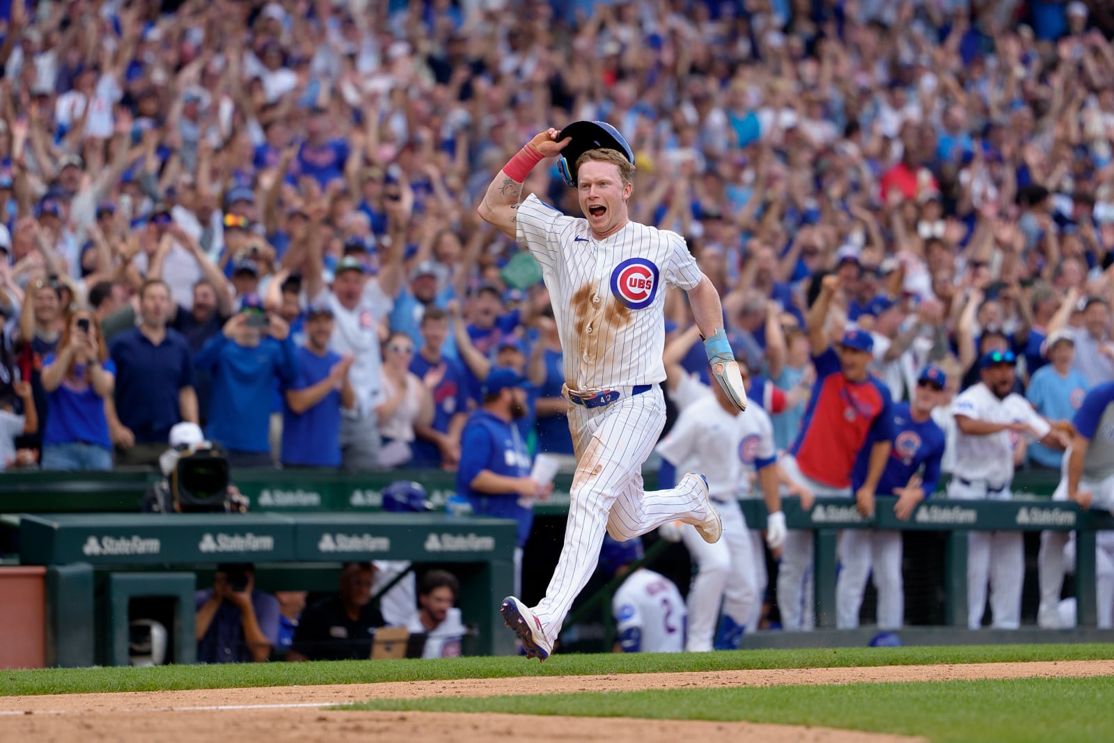 A Chicago Cubs baseball player in a pinstriped uniform runs across the field with his helmet raised, celebrating in front of a cheering crowd.