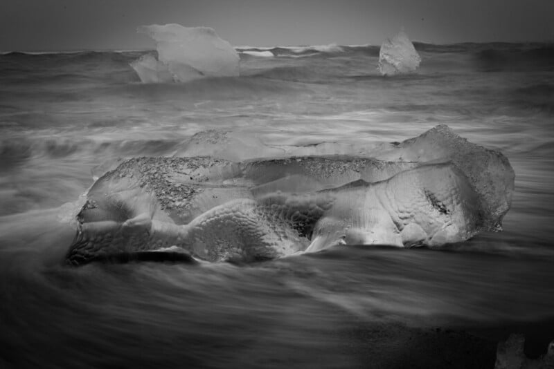 A large, translucent chunk of ice rests on a dark, wet beach as ocean waves wash over it, with two smaller icebergs in the distance.