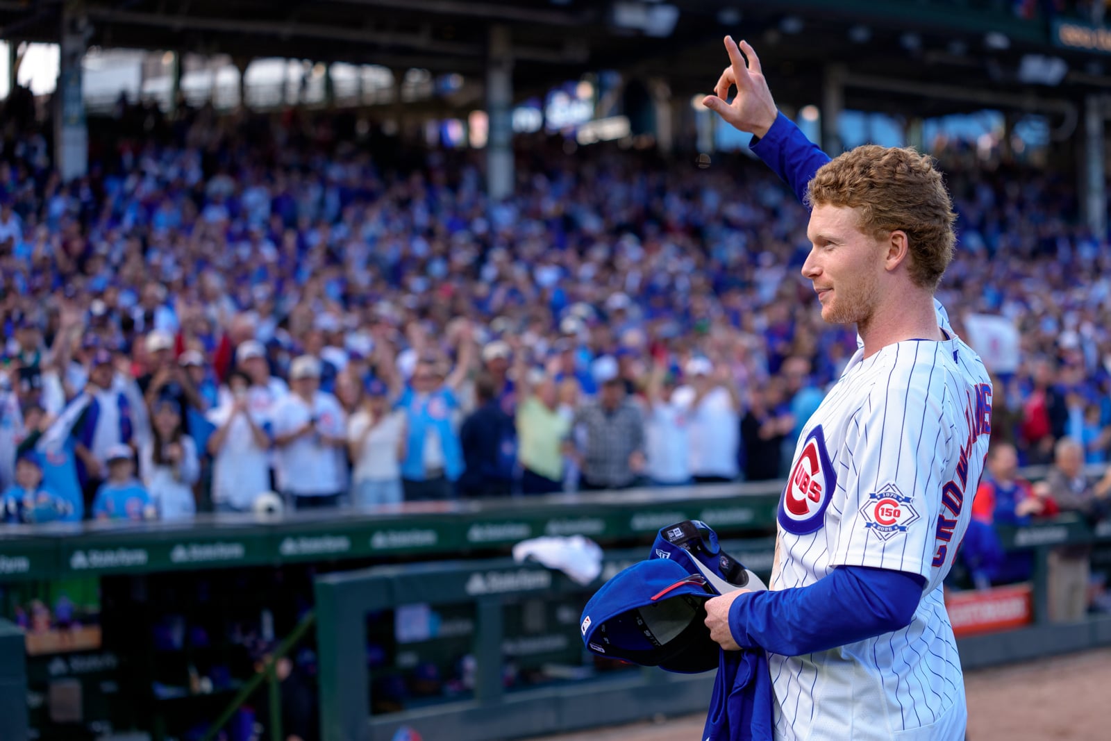 A baseball player in a Chicago Cubs uniform stands on the field, holding his cap and waving to a cheering crowd in the stands.
