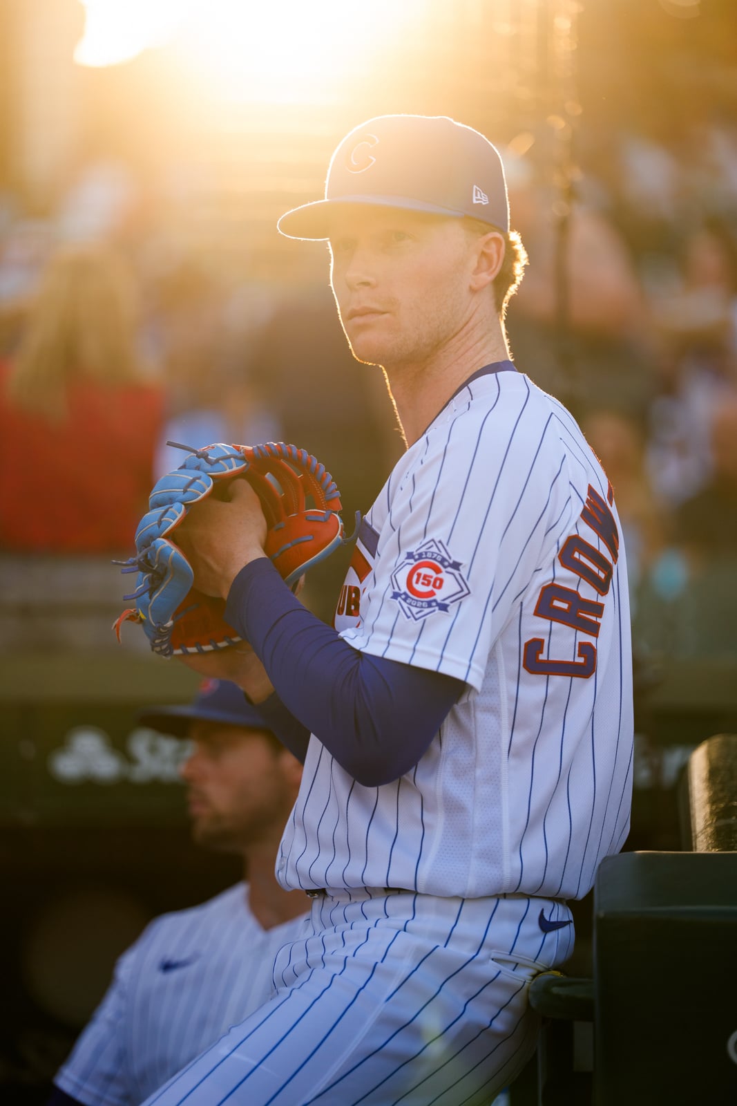 A baseball player in a Chicago Cubs uniform sits in the dugout during a sunset, holding a blue and red glove.