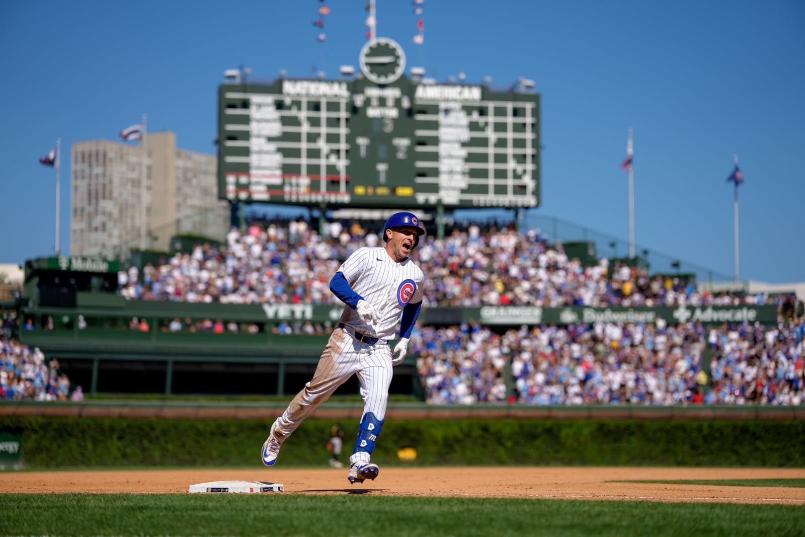 A Chicago Cubs baseball player in a pinstriped uniform runs across the field toward a base with a large scoreboard in the background.