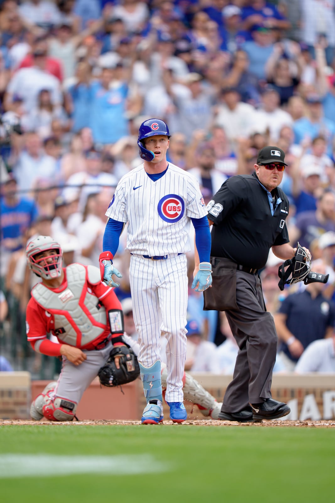 A Chicago Cubs baseball player stands at home plate after a hit, while the opposing catcher kneels behind him and an umpire stands to the right.