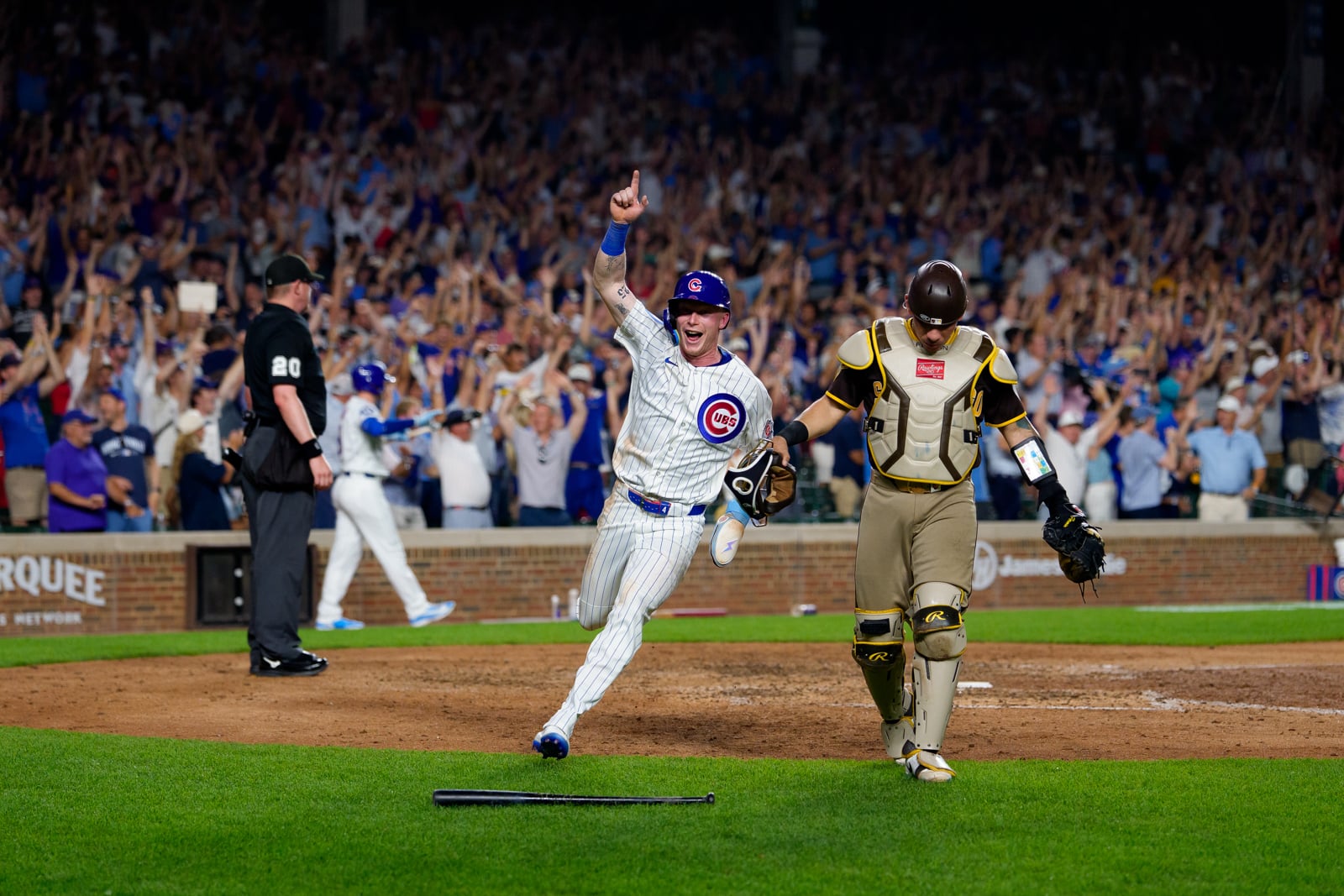 A Chicago Cubs player celebrates with his arm raised while running past a San Diego Padres catcher on a baseball field in front of a cheering crowd.