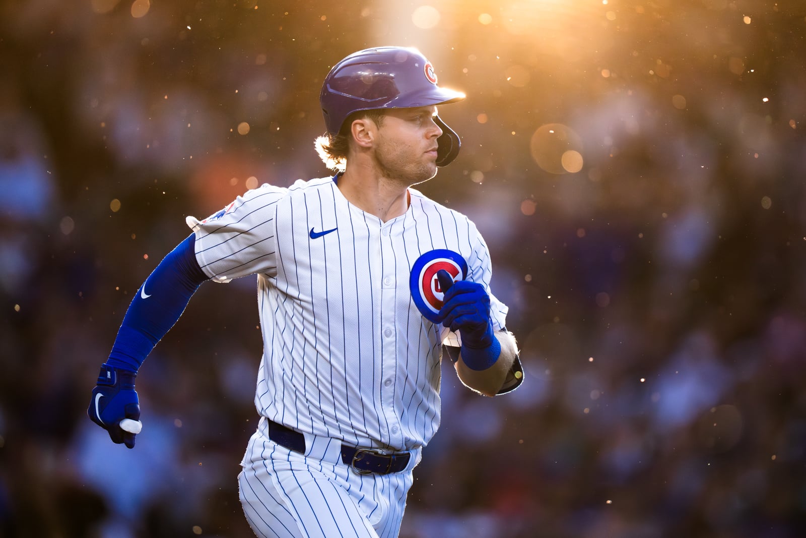 A Chicago Cubs baseball player in a pinstriped uniform runs across the field during a sunny, backlit game.