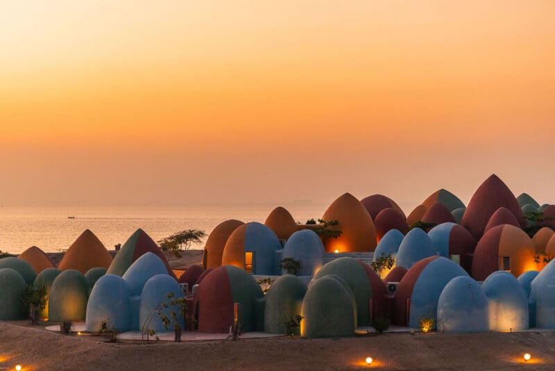 A cluster of colorful, dome-shaped buildings sits along a coastline at sunset, with the ocean visible in the background.