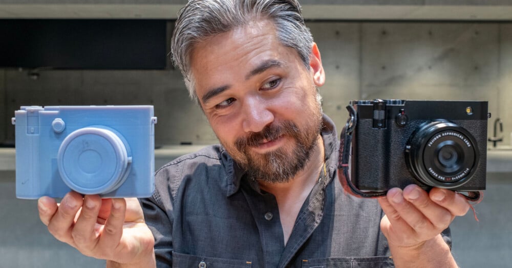A man with graying hair and a beard holds a light blue 3D-printed camera model in his left hand and a black digital camera in his right hand.