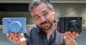 A man with graying hair and a beard holds a light blue 3D-printed camera model in his left hand and a black digital camera in his right hand.