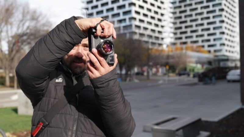 A man with a beard wearing a black puffer jacket holds a camera up to his face to take a picture outdoors.