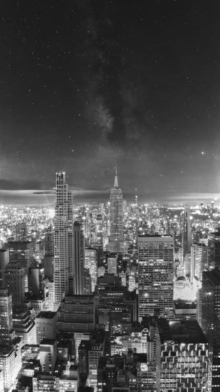 A black and white aerial view of a dense city skyline at night under a starry sky with a visible Milky Way.