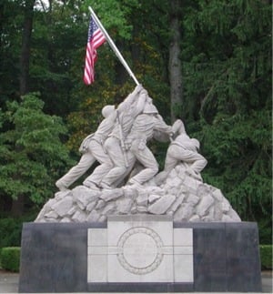 A stone sculpture of several soldiers raising an American flag on a rocky mound, set against a background of green trees.