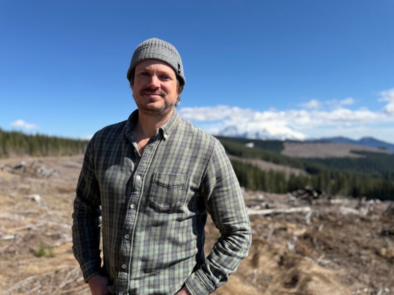 A man with a mustache and a grey beanie wears a plaid button-down shirt while standing in a cleared forest area with a snow-capped mountain in the distance.