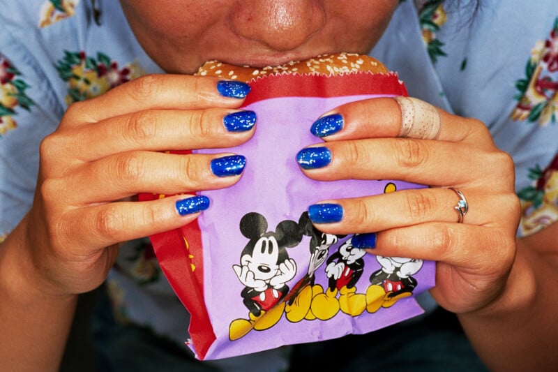 A person with blue glittery nail polish holds a burger in a purple Mickey Mouse wrapper while taking a bite.