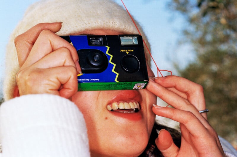 A person wearing a cream-colored knit hat holds a disposable camera with a Walt Disney Company logo up to their face, partially obscuring their eyes while smiling.