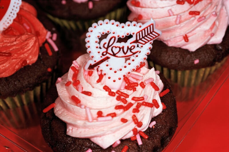 Chocolate cupcakes topped with pink frosting and red sprinkles, featuring a white heart-shaped "Love" topper with an arrow.