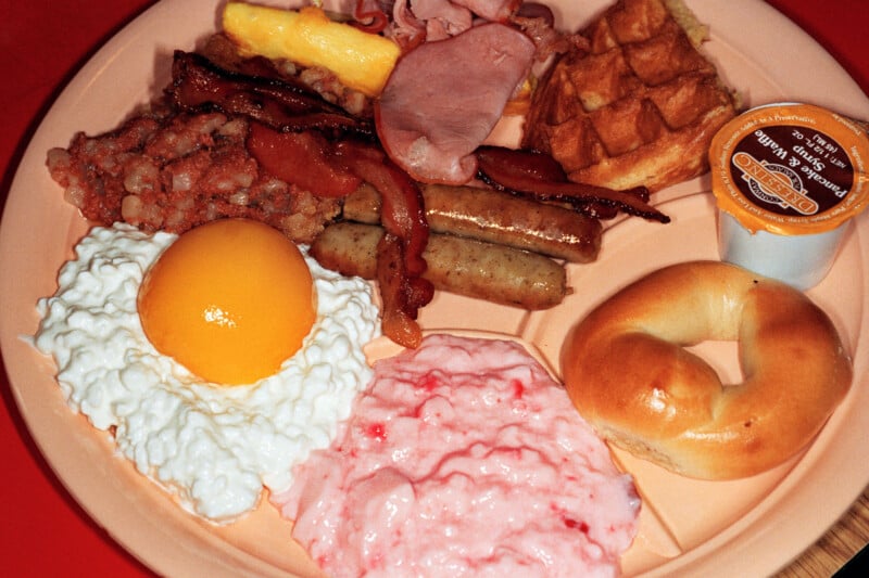 A divided plate holds a breakfast meal featuring a waffle, sausage links, bacon, ham, corned beef hash, a bagel, cottage cheese with a peach half, and pink yogurt.
