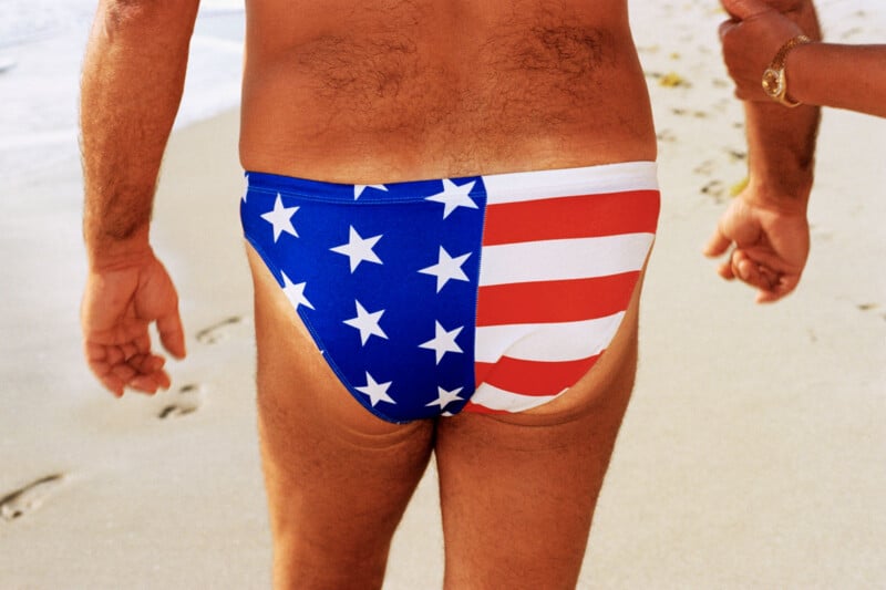 A person walks on a sandy beach wearing American flag-patterned swim briefs, with another person's arm and gold watch visible on the right.