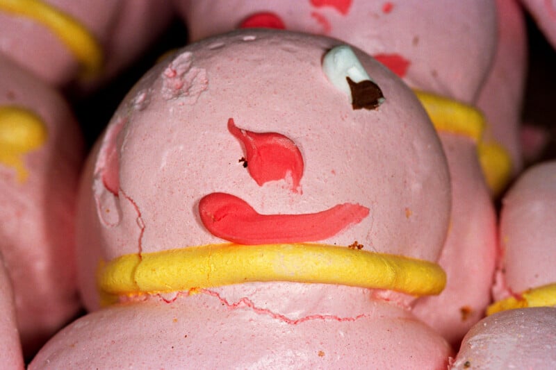 A close-up of a pink, snowman-shaped meringue cookie decorated with a yellow icing ring, a red icing nose and mouth, and a small white and brown eye.