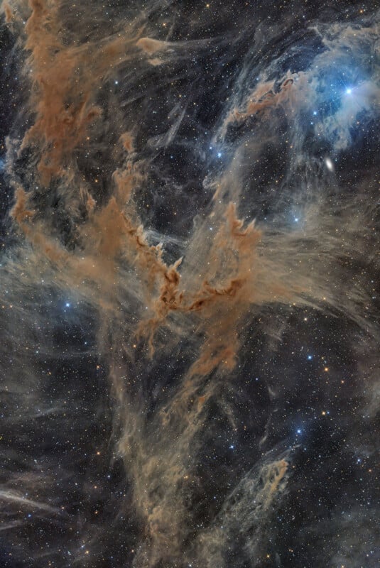 A vast, wispy nebula of brown dust clouds and faint blue gas filaments set against a dense field of distant stars.