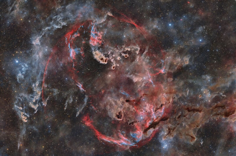 A vibrant nebula features glowing red and blue filaments of gas swirling against a dense, dark background of distant stars.