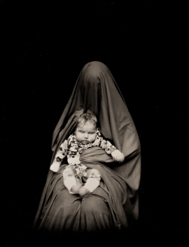 A baby in a patterned outfit sits on the lap of a person completely covered by a dark, flowing fabric against a black background.