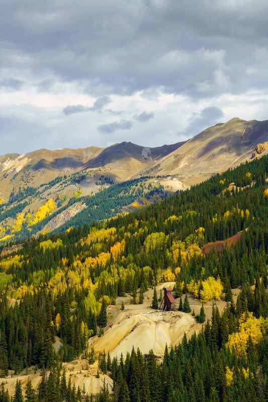 A rustic wooden mining structure stands on a barren, light-colored slope surrounded by a dense forest of evergreen and yellow autumn trees beneath a cloudy sky.