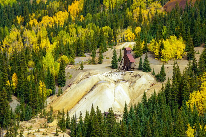 An old, weathered wooden mining headframe stands on a barren, light-colored tailings pile surrounded by a dense forest of evergreen and yellow autumn aspen trees.