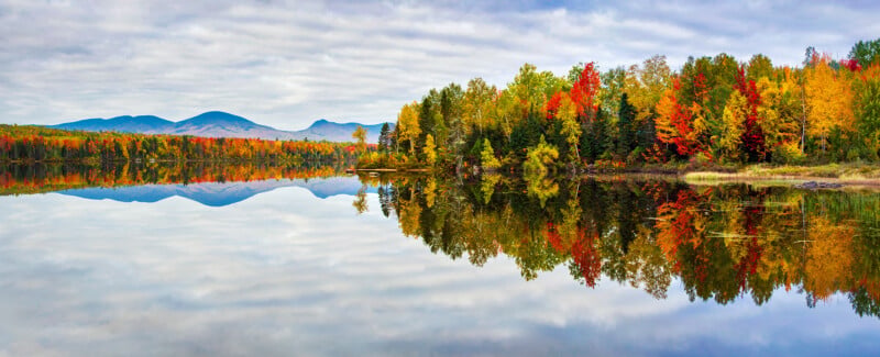 A calm lake reflects a vibrant autumn forest and distant mountains under a cloudy sky.