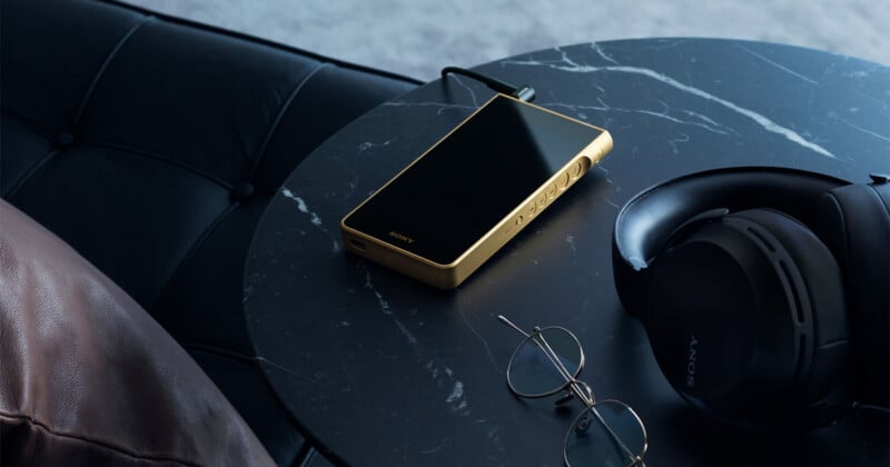 A gold-colored Sony digital music player with a black screen sits on a dark marble table next to a pair of black Sony headphones and wire-rimmed glasses.