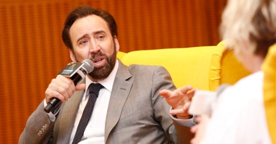 Nicolas Cage sits in a yellow chair, holding a microphone and gesturing while speaking to someone off-camera.