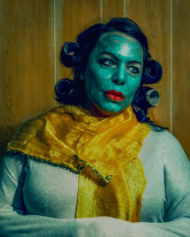 A woman with blue-tinted skin and bright red lipstick wears hair rollers and a yellow scarf against a wood-paneled wall.