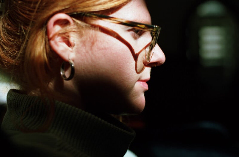 A profile view of a person with reddish hair wearing glasses, a hoop earring, and a green turtleneck sweater.