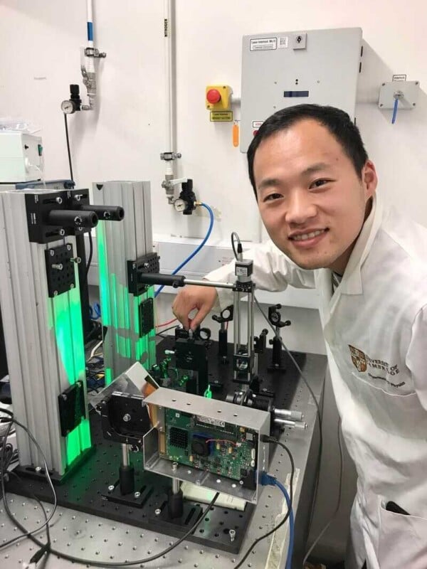 A smiling man in a white lab coat works with optical equipment and a circuit board on an experimental table in a laboratory.