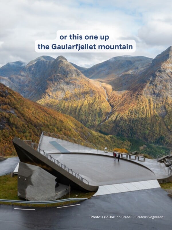 A modern concrete viewing platform with a metal railing overlooks a deep valley surrounded by steep, autumn-colored mountains.