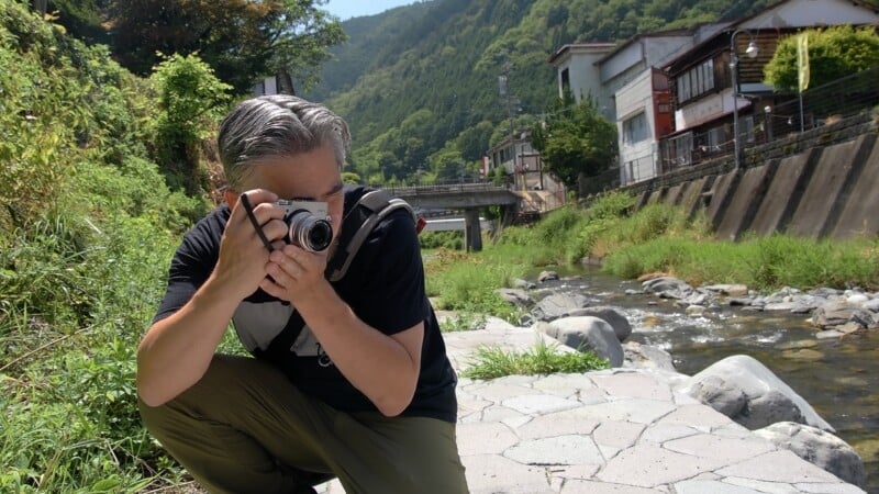A man with gray hair crouches by a rocky stream, holding a camera up to his eyes to take a picture.