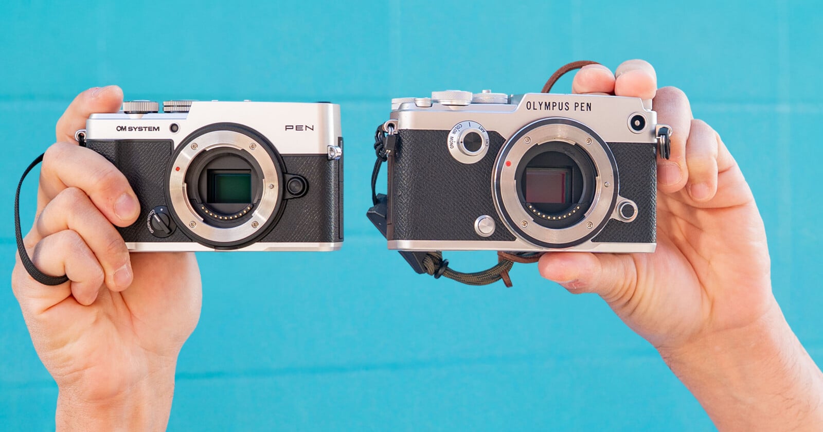 Two hands hold two silver and black mirrorless cameras against a blue tiled background; the left camera is labeled "OM SYSTEM" and "PEN," and the right is labeled "OLYMPUS PEN.