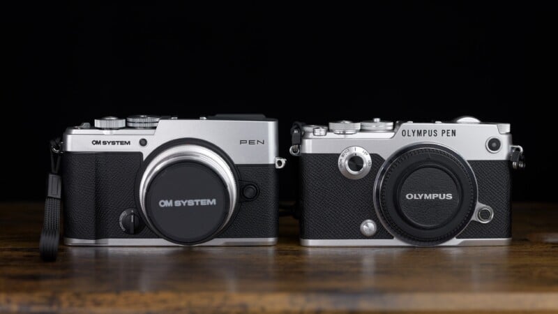 Two silver and black mirrorless cameras sit side-by-side on a wooden surface against a black background.